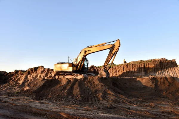 Excavator working on earthmoving. Backhoe digs ground in sand quarry on blue sky background. Construction machinery for excavation, loading, lifting and hauling of cargo on job sites