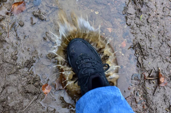 Water splash from shoes. Men's feet in hiking shoes steps into a puddle. Rain footwear for man or woman. Trekker boots for for cold and weather hike. Pair of waterproof travel shoe in blue jeans