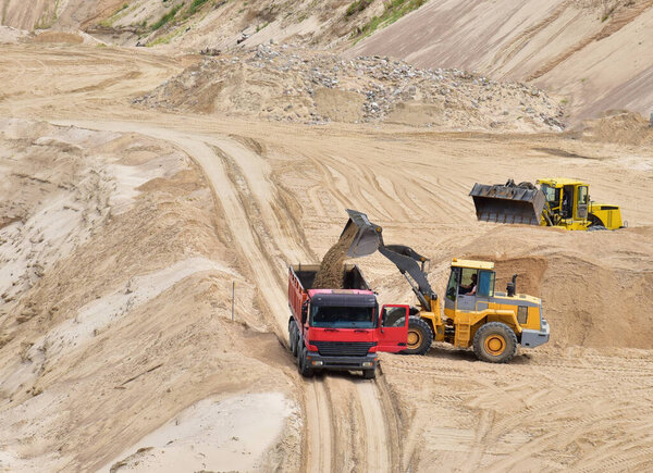 Wheel loader loading sand into heavy dump truck at the opencast mining quarry. Dump truck transports sand in open pit mine. Quarry in which sand and gravel is excavated from the ground.