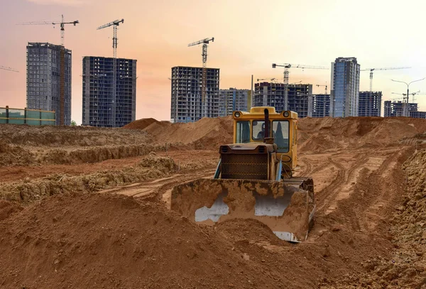 Dozer on earthmoving at construction site on sunset background. Construction machinery and equipment on groundwork. Bulldozer leveling ground for new road construction. Tower cranes in action