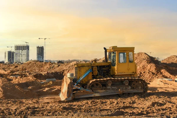 Dozer on earthmoving at construction site on sunset background. Construction machinery and equipment on groundwork. Bulldozer leveling ground for new road construction. Tower cranes in action