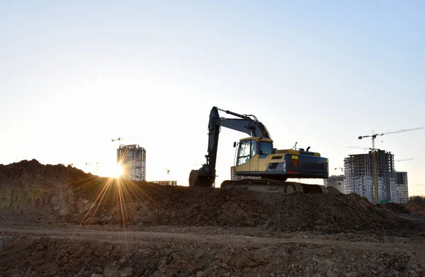 Excavator during earthmoving at construction site on sunset background. Backhoe digs ground for underground sewage and water pipes. onstruction machinery for excavating