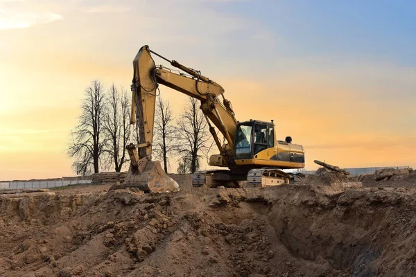 Excavator on earthworks at construction site on sunset background. Digging a foundation pit and a trench for laying sewer pipes.  Renovation and construction of a residential multi-storey building