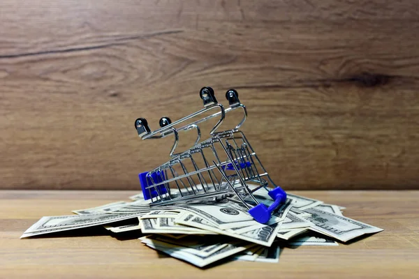 Supermarket basket lies on a heap with American dollars. Shopping cart trolley at Hundred Dollar Bills. Lots of money for shopping. The cost of food and clothing in the store. Social problem