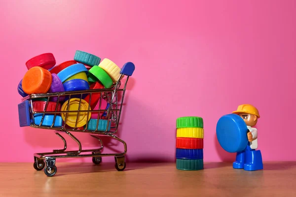 Multicolored Plastic bottle caps in shopping trolley on pink background. Cap material is recyclable. Lids from plastic bottles ror recycling