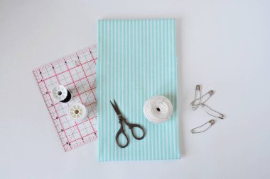 Çizgili pamuk kumaş, küçük makas, iplikler, iğne, örtü cetveli ve beyaz üzerine iğneler.