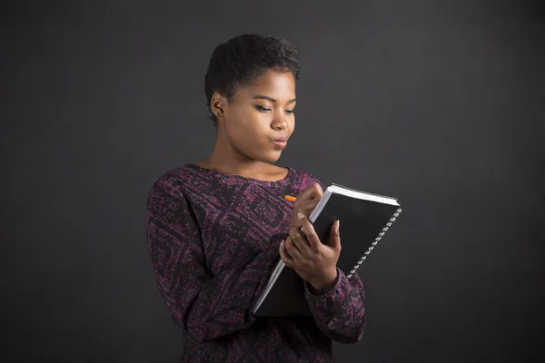 African American woman writing in book diary on blackboard background — Stockfoto