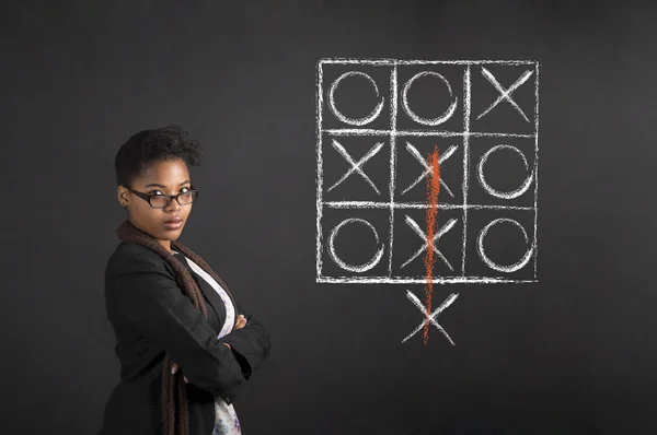 South African or African American woman teacher or student with arms folded tic tac toe diagram on chalk black board background — Stock Photo, Image