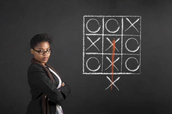 South African or African American woman teacher or student with arms folded tic tac toe diagram on chalk black board background Stock Picture