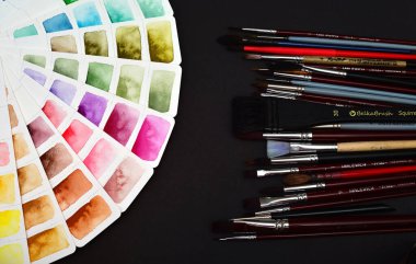 Beyaz kağıt üzerinde suluboya palet, bir dizi fırçanın yanında serpilmiş. Siyah bir arkaplanda.