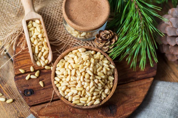 Los piñones en la escudilla, el cono de pino, la rama, el frasco de cristal en la tabla de cortar. Bodegón de ingredientes naturales, vista superior. Imagen de stock