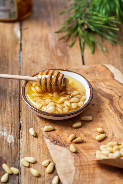 A plate with honey and pine nuts, a honey spoon with transparent honey dripping from it. Light wooden table background, behind a pine branch.