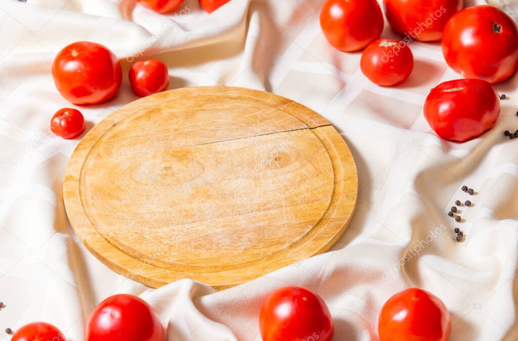 Round wooden cutting board, in around the edges are red ripe tomatoes on a tablecloth. Side view