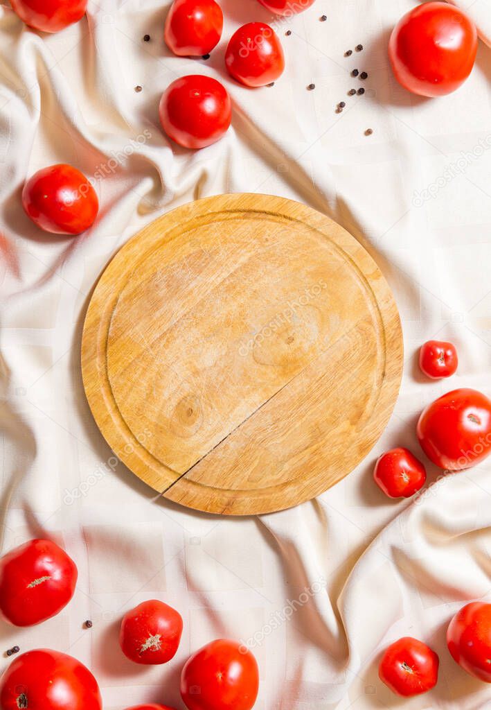 Round wooden cutting board, in around the edges are red ripe tomatoes on a tablecloth. Vertical