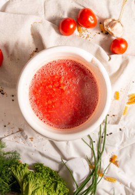 Etrafında taze sebzeler olan bir kase domates çorbası..