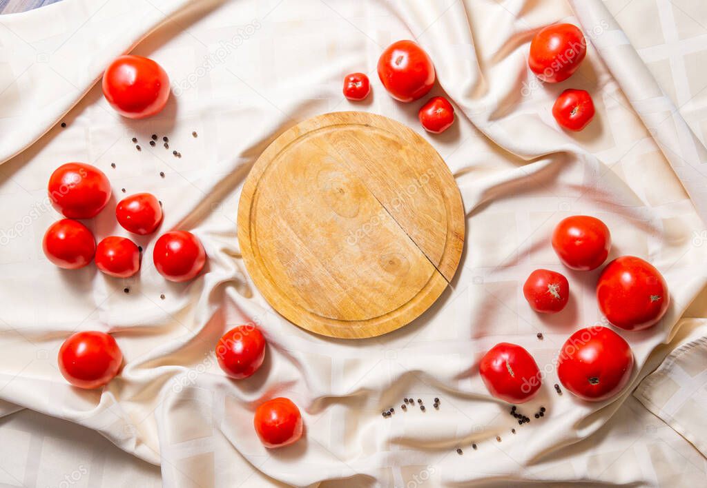Round wooden cutting board, in around the edges are red ripe tomatoes on a tablecloth.