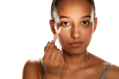 Genç, koyu tenli bir kadın, beyaz arka planda alçak göz kapaklarına kapatıcı sürüyor.