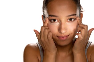 Genç, koyu tenli bir kadın, beyaz arka planda alçak göz kapaklarına kapatıcı sürüyor.