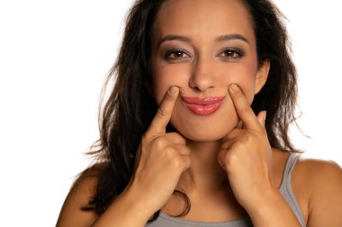 Genç esmer tenli kadın, beyaz arka planda parmaklarıyla gülümsemeye zorluyor.