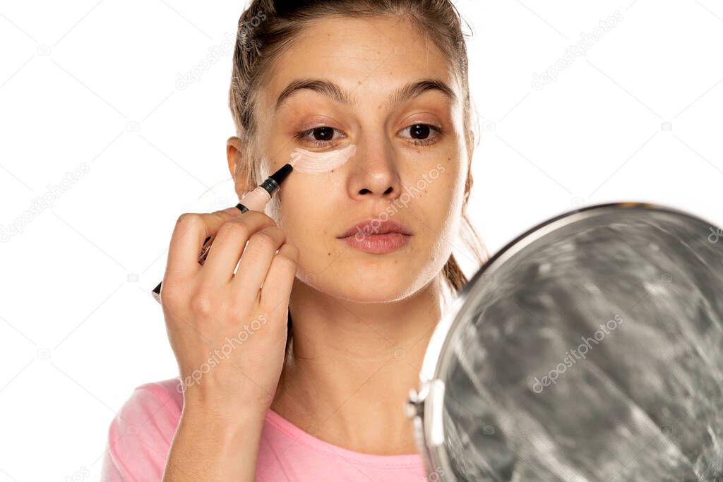 Portrait of young beautiful woman applying concealer with appicator on white background