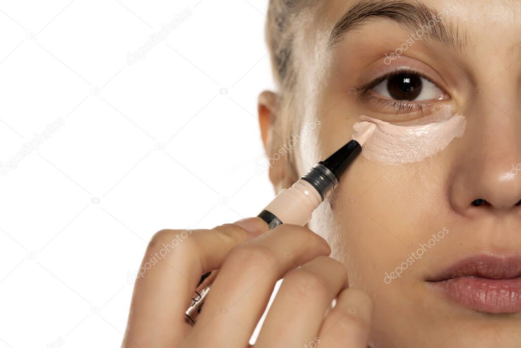 Portrait of young beautiful woman applying concealer with brush on white background