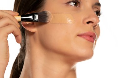 Genç bir kadın, beyaz arka planda yüzünde fırçayla sıvı fondöten uyguluyor.