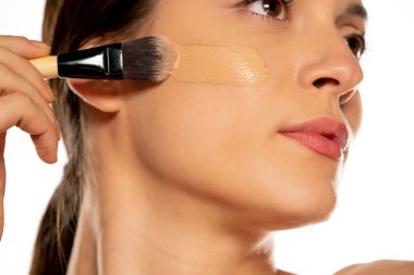 Yüzünde fırçayla sıvı fondöten süren genç bir kadın.