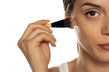 Yüzünde fırçayla sıvı fondöten süren genç bir kadın.