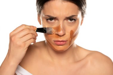 Genç, gergin bir kadın beyaz arka planda yüzüne üç farklı tonda sıvı fondöten sürüyor.