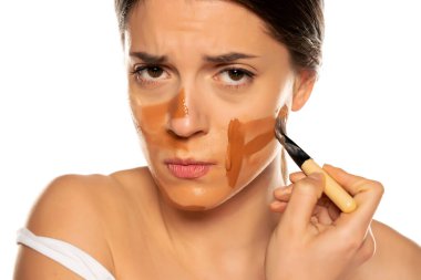Genç, gergin bir kadın beyaz arka planda yüzüne üç farklı tonda sıvı fondöten sürüyor.