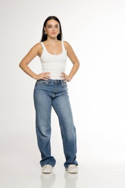 Studio shot of young woman with long dark hair wearing white tank top, blue jeans and sneakers posing full length with hands on hips on white background