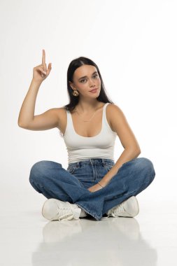 Studio shot of young woman with long dark hair wearing white tank top, blue jeans and sneakers sitting cross legged and pointing up on white background