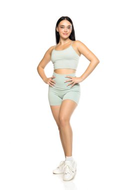 Studio shot of full length young woman in mint green activewear and white sneakers, standing with hands on hips and confident expression on white background