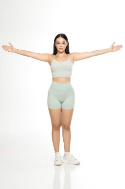 Nane yeşili aktif giyimli, beyaz spor ayakkabılı, kolları beyaz arka planda anatomik veya fitness pozu için yatay olarak açılmış genç bir kadının stüdyo fotoğrafı.