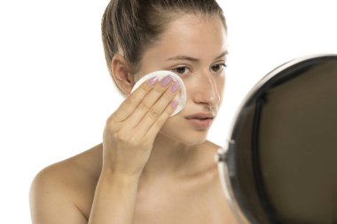 Studio shot of young woman with no makeup cleaning face with cotton pad in mirror on white background