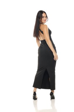 Sırtında siyah elbisesi olan, sırtüstü gülümseyen, arkasını kollayan, beyaz arka plandaki kameraya bakan esmer bir kadının stüdyo fotoğrafı.