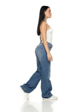 Uzun siyah saçlı, beyaz kolsuz bluzlu, mavi kot pantolonlu ve spor ayakkabılı bir kadının stüdyo fotoğrafı. Bir bacağını beyaz arka planda kaldırıyor.