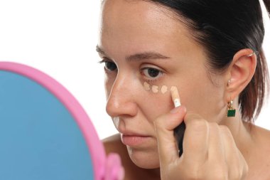 Siyah saçlı ve makyajsız doğal bir kadının beyaz arka planda aynaya bakarken gözlerinin altına kapatıcı sürdüğü bir fotoğraf.