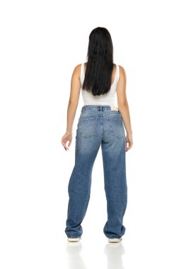 Uzun siyah saçlı, beyaz kolsuz bluzlu, kot pantolonlu ve spor ayakkabılı, beyaz arka planda dikiz aynasından bakan genç bir kadının stüdyo fotoğrafı.