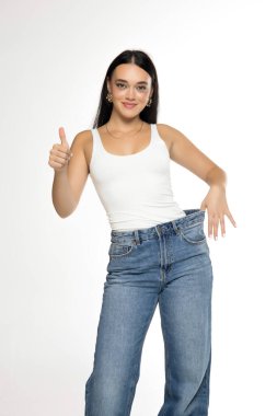 Stüdyo resmi, uzun siyah saçlı, beyaz atlet giyen, bel kemeri takan ve beyaz arka planda baş parmaklarını gösteren açık mavi kotlu genç bir kadın.