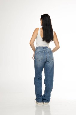 Uzun siyah saçlı, beyaz kolsuz bluzlu, kot pantolonlu ve spor ayakkabılı, beyaz arka planda dikiz aynasından bakan genç bir kadının stüdyo fotoğrafı.