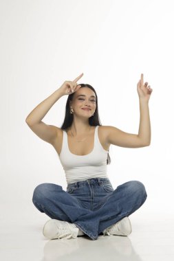 Uzun siyah saçlı, beyaz kolsuz bluzlu, kot pantolonlu, spor ayakkabılı çapraz bacaklı ve iki eli de beyaz arka planda olan genç bir kadının stüdyo fotoğrafı.