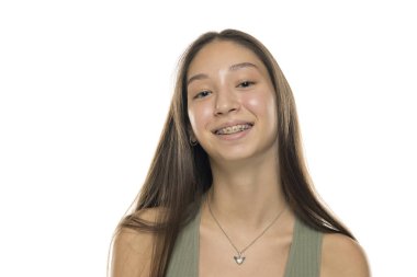 Uzun kahverengi saçlı, yeşil kolsuz tişörtlü, diş telleriyle gülümseyen beyaz arka planda genç bir kadının stüdyo fotoğrafı.