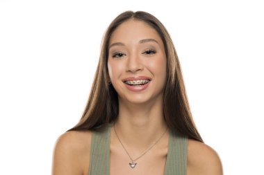 Uzun kahverengi saçlı, yeşil kolsuz tişörtlü, diş telleriyle gülümseyen beyaz arka planda genç bir kadının stüdyo fotoğrafı.