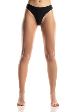 Bir kadının bacakları ve alt vücudu ön planda, siyah külot giymiş, pürüzsüz ciltli yalınayak duran, beyaz bir arka planda stüdyo çekimi yapan bir kadın..