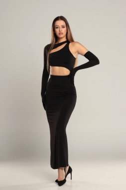 Uzun boylu, esmer bir kadının zarif, siyah, asimetrik bir gece elbisesi ve gri arka planda uzun eldivenler giydiği bir fotoğraf stüdyosu..