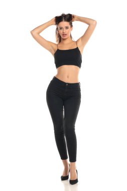 Uzun boylu, esmer, uzun saçlı, siyah bir bluz giyen, dar kotlu ve yüksek topuklu giyen bir kadının beyaz arka planda elleri ile poz verdiği bir stüdyo resmi.