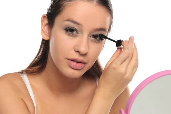 Молодая женщина наносит тушь, глядя в зеркало, студия снимается на белом фоне.