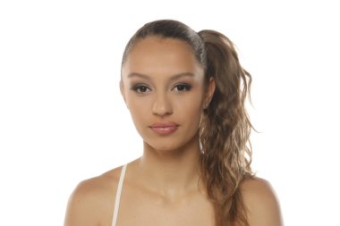 Stüdyoda, beyaz arka planda beyaz kayışlı bir bluz giyen, uzun saçlı, kahverengi dalgalı saçlı genç bir kadın görülüyor.