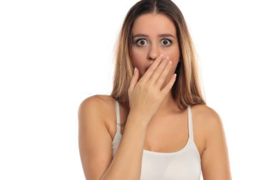 Uzun kahverengi saçlı ve makyajlı, beyaz bir bluz giyen ve beyaz bir arka planda elini kullanarak ağzını kapatan şaşırmış bir kadının stüdyo fotoğrafı..